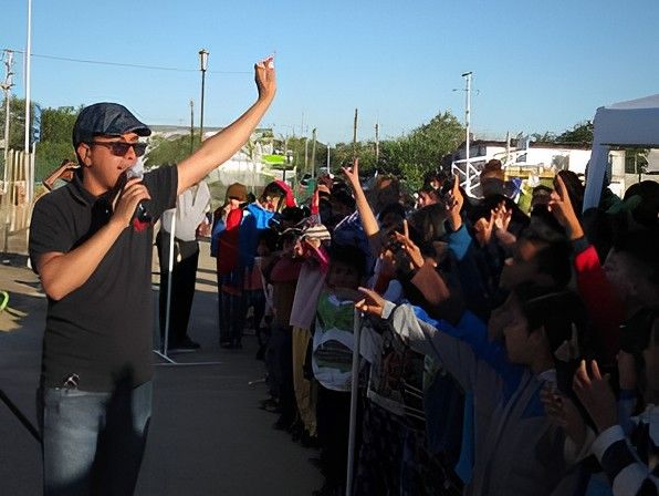 Man speaking into microphone, arm raised, addressing a crowd of children outdoors.