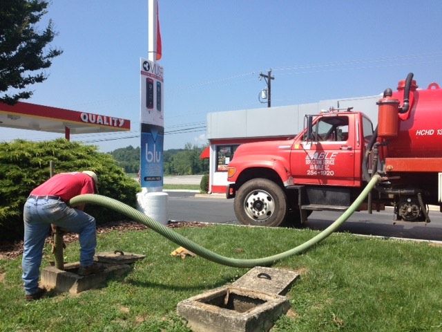 Man in red shirt connecting hose to a septic tank; red truck at a gas station.