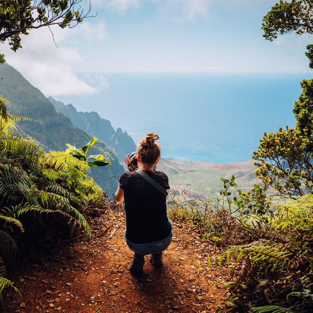 Woman on a hiking trail looks out over a mountain and ocean vista, holding a camera. Green foliage surrounds the path.