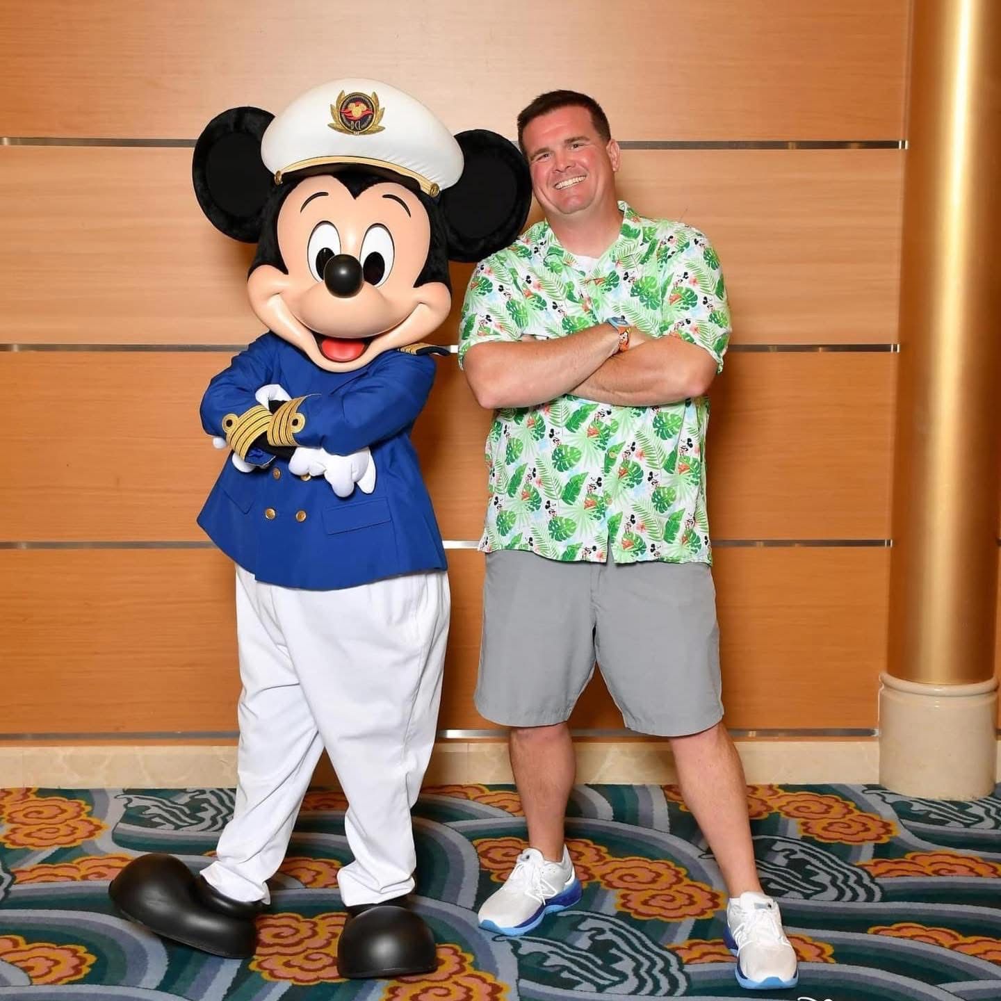 Mickey Mouse in sailor attire poses with a smiling man wearing a Hawaiian shirt and gray shorts.