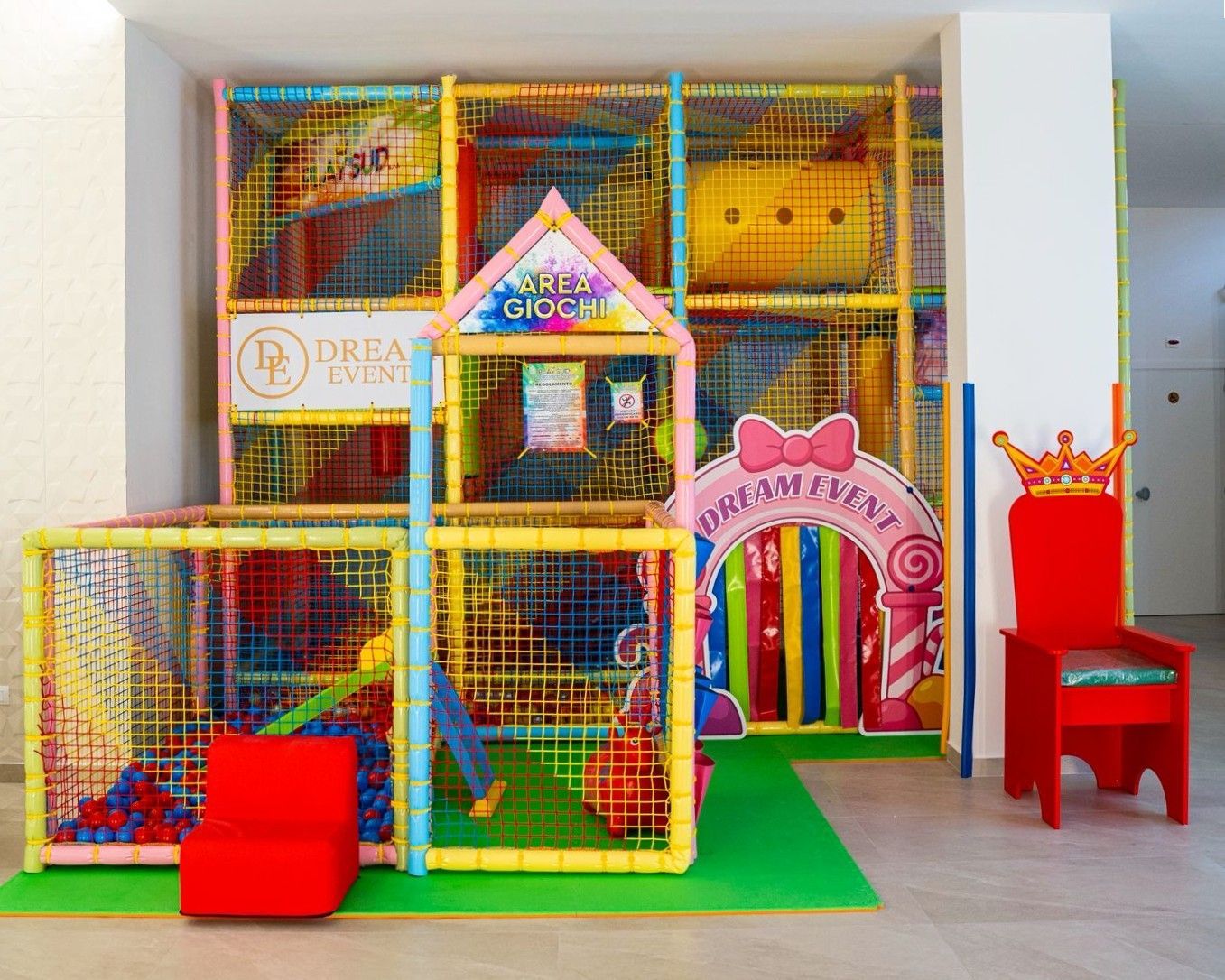 Area giochi al coperto con una struttura per arrampicarsi, una piscina di palline, uno scivolo e un trono rosso