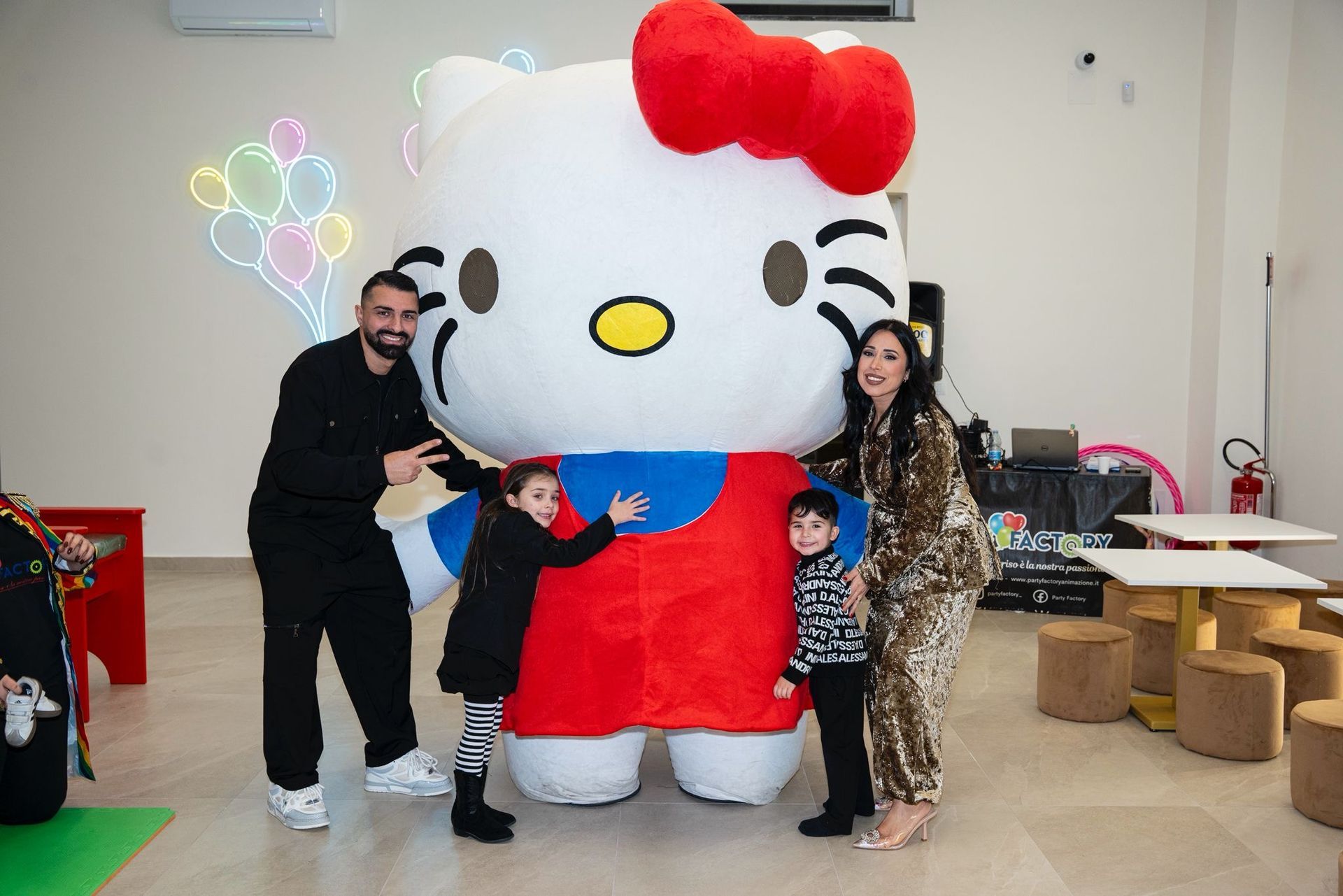 Una famiglia posa con una grande mascotte di Hello Kitty in uno spazio per eventi decorato con palloncini festivi.