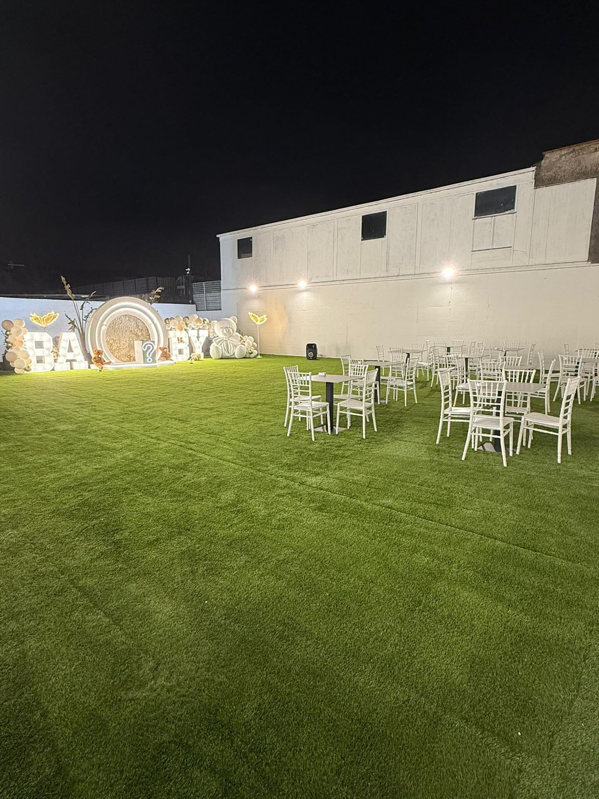 Un evento serale all'aperto su un prato con tavoli e sedie bianchi e un cartello decorativo con la scritta 