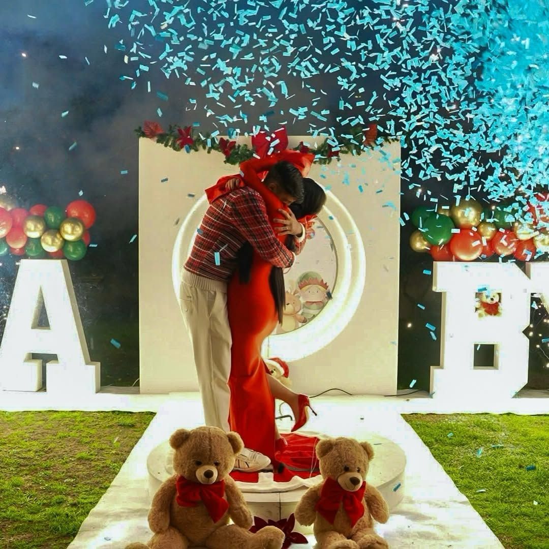 Una coppia si abbraccia tra coriandoli blu con grandi lettere, palloncini e due orsacchiotti su una piattaforma.