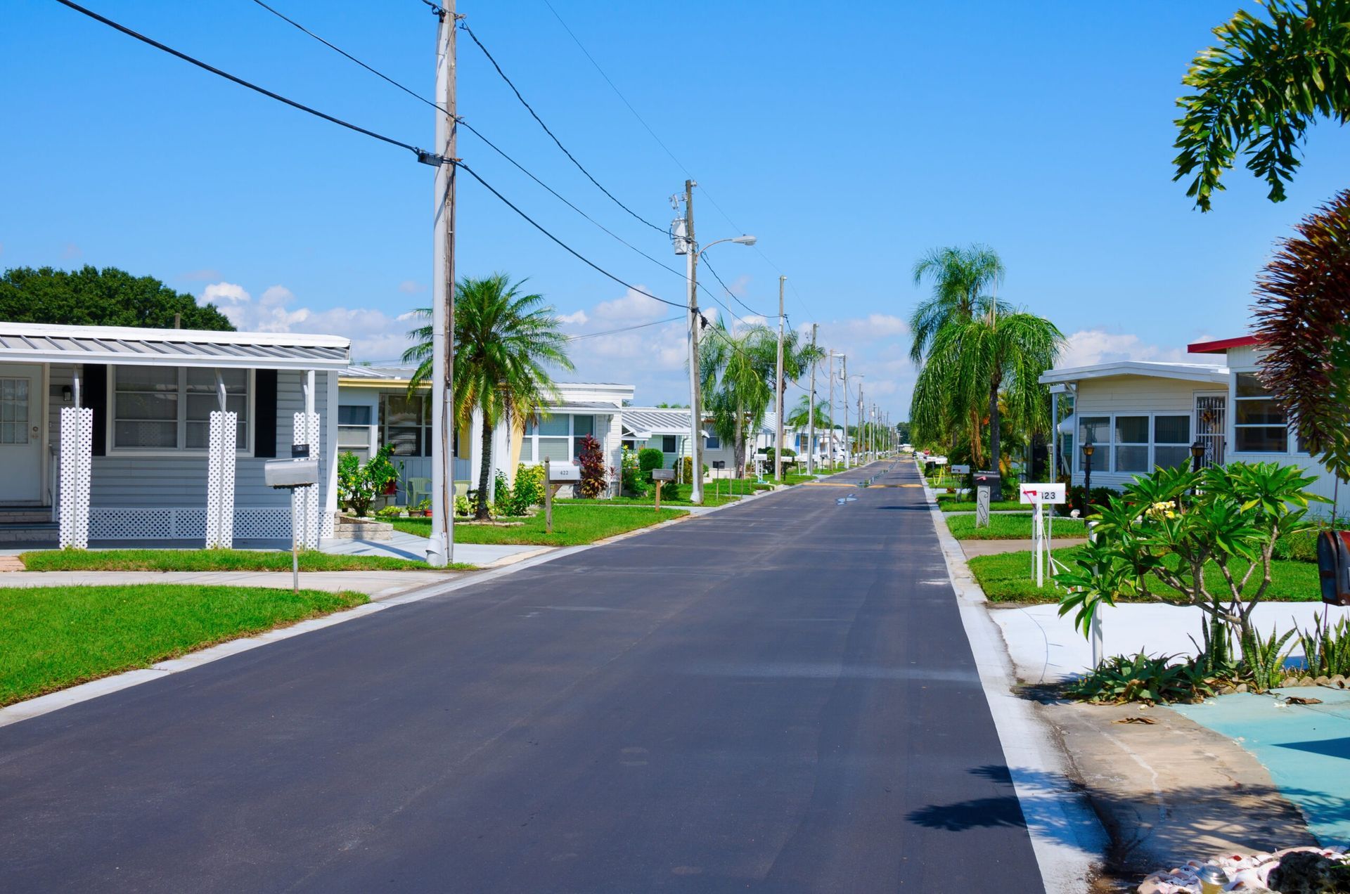 Street in a mobile home park under a blue sky. White homes with green lawns line the road.