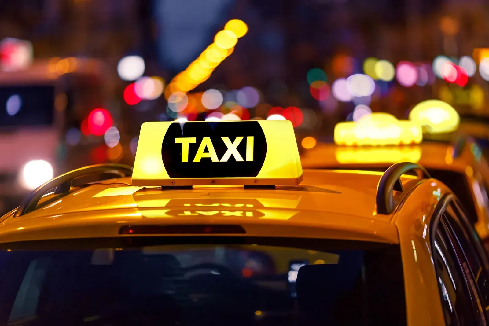Yellow taxi cab at night, lit “TAXI” sign atop, blurred city lights in background.