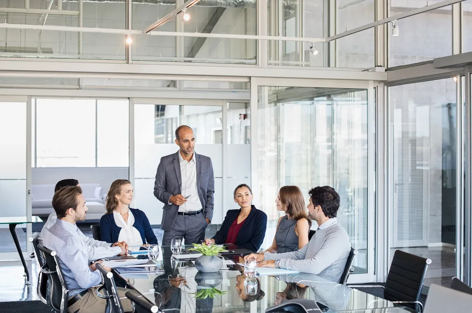 Business meeting: A bald man presents at a conference table with six colleagues in a bright office.
