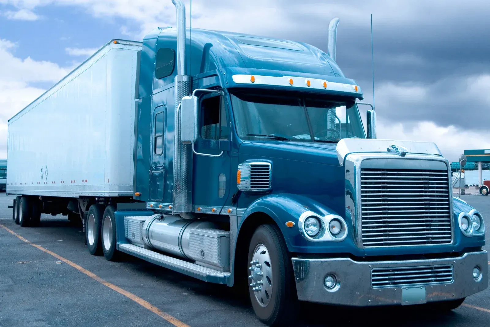 Blue semi-truck with a white trailer parked outdoors under a cloudy sky.