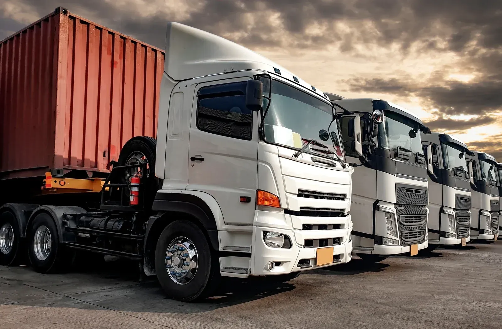 Semi-trucks parked in a row, one with a red container, under a cloudy sky.