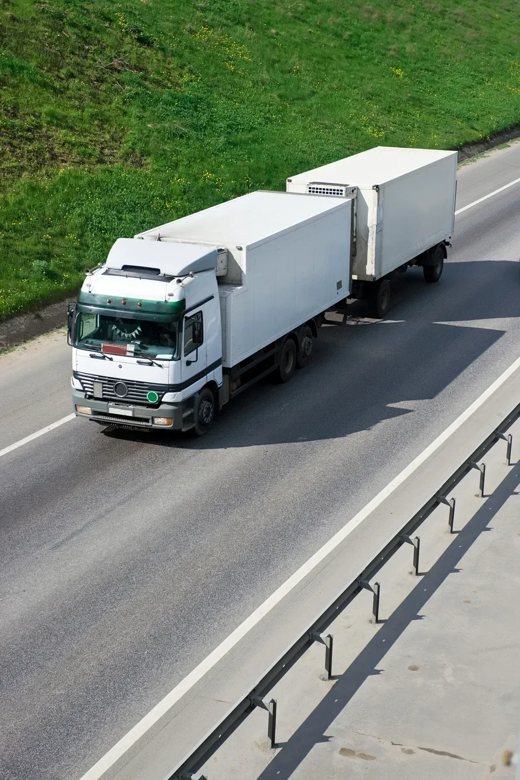 White semi-truck with two trailers driving on a highway, green hill in the background.