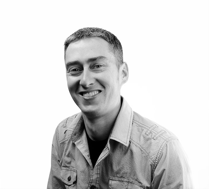 A man in a denim shirt is smiling in a black and white photo.