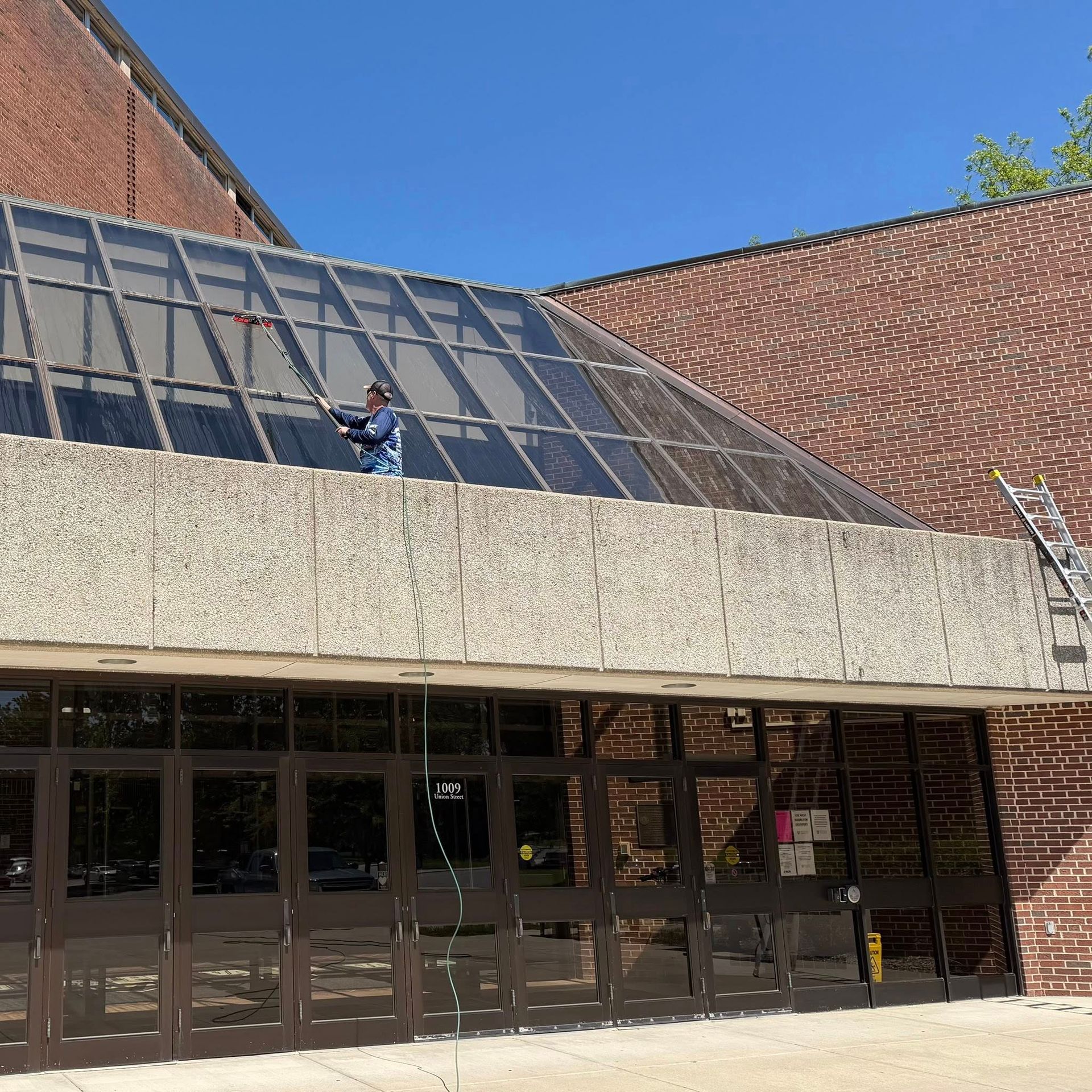 Brick building entrance with large glass windows, concrete overhang, and a worker on the sloped roof