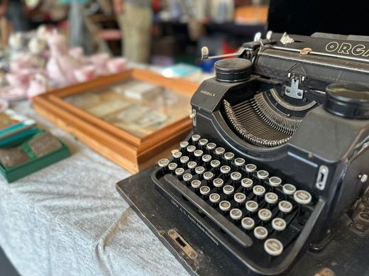 Eine antike schwarze Schreibmaschine steht auf einem Tisch auf einem Antiquitätenmarkt; im Hintergrund sind gerahmte Gegenstände und Glaswaren zu sehen.