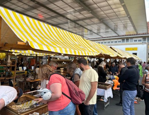 Ein geschäftiger Marktstand im Freien unter einer gelb-weiß gestreiften Markise, an dem Käufer Obst, Gemüse und andere Waren durchstöbern.