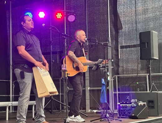 Zwei Musiker treten auf einer Freilichtbühne unter violettem Licht auf, einer singt mit Gitarre, der andere spielt Cajón.