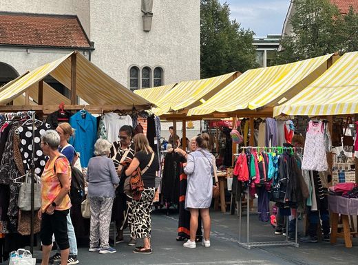 Freiluftmarkt mit Käufern, die unter gelb-weiß gestreiften Markisen an Bekleidungsständen stöbern.