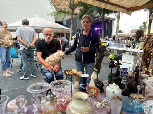 Marktstand im Freien mit Glaswaren und Figuren, zwei Personen stöbern unter einer Markise.