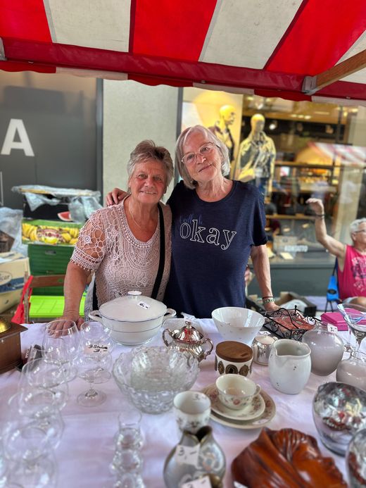 Zwei lächelnde Menschen hinter einem Markttisch voller Glaswaren und Keramik unter einem roten Baldachin.