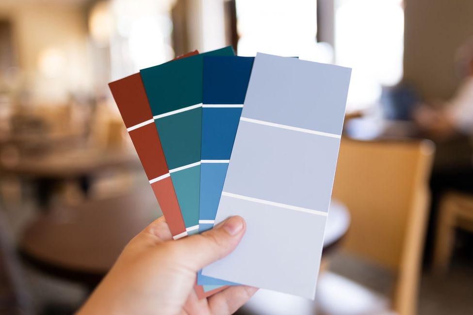 Hand holding three color swatch cards with teal, red, and light blue panels in a blurred indoor setting