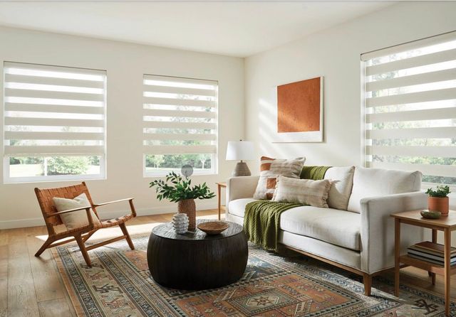 Bright living room with white sofa, wooden chair, round coffee table, patterned rug, and large windows with blinds