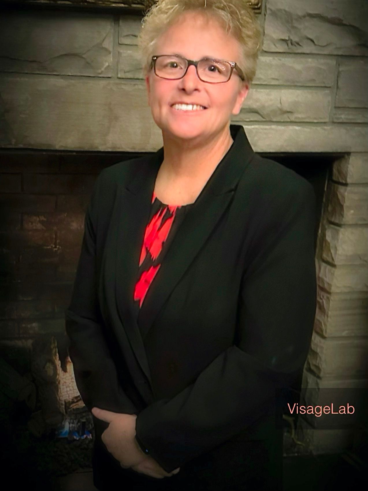 A woman wearing glasses and a black jacket is standing in front of a fireplace.