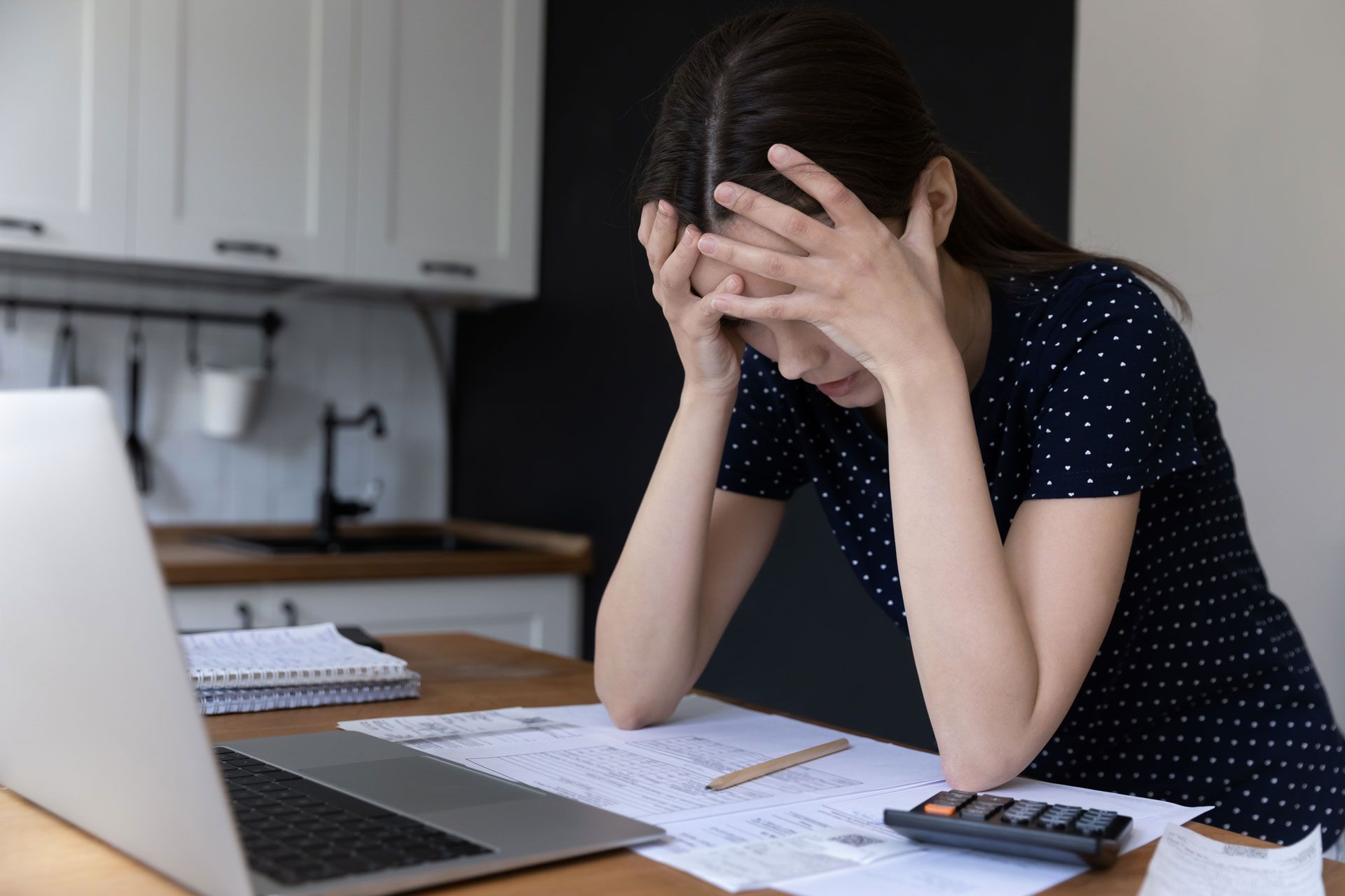Woman with head in hands, stressed over bills and laptop at kitchen table.