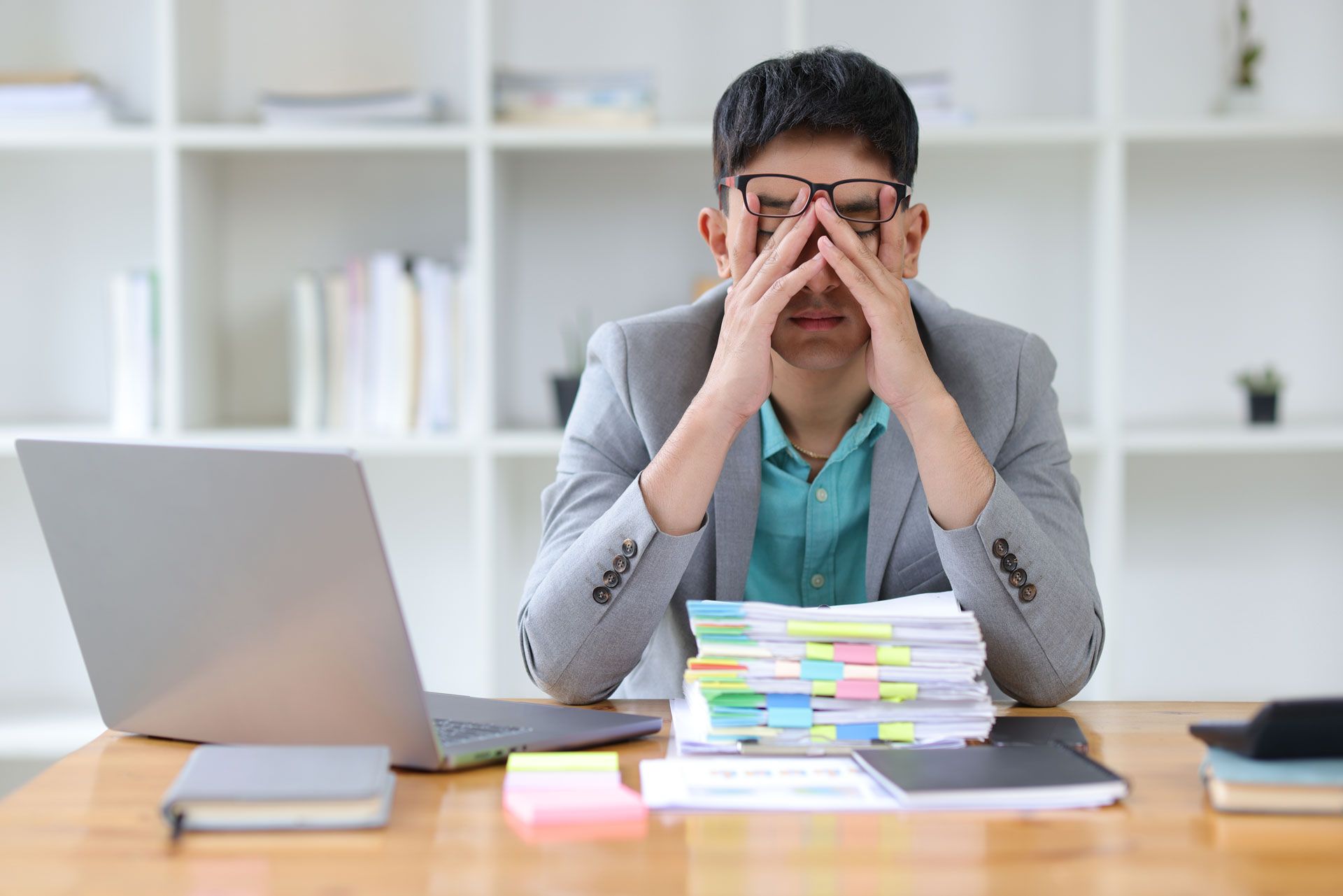 Man with glasses at desk, hands on face, looking stressed. Laptop, documents, and notepad are visible.