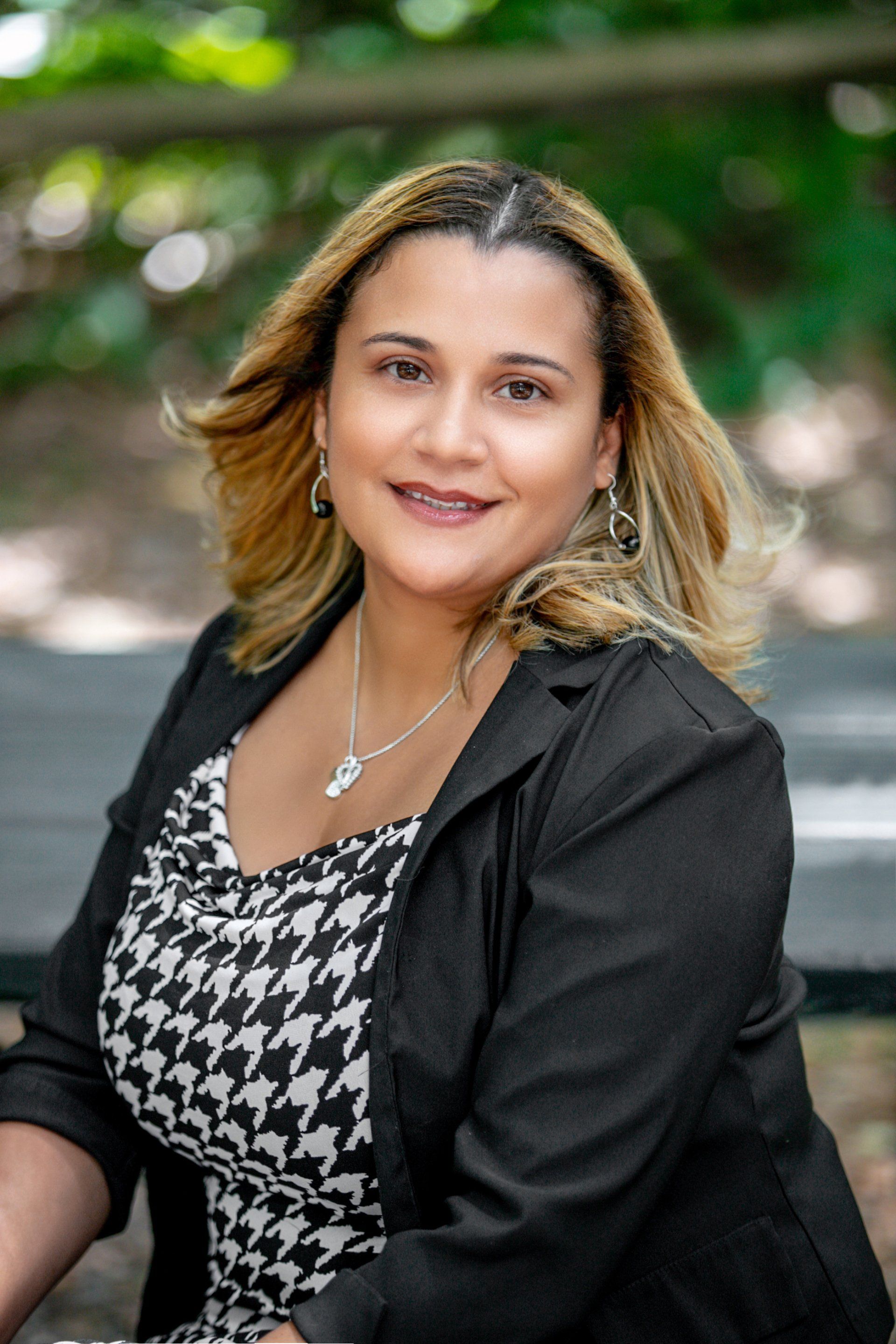 A woman is sitting on a park bench wearing a black jacket and a black and white dress.