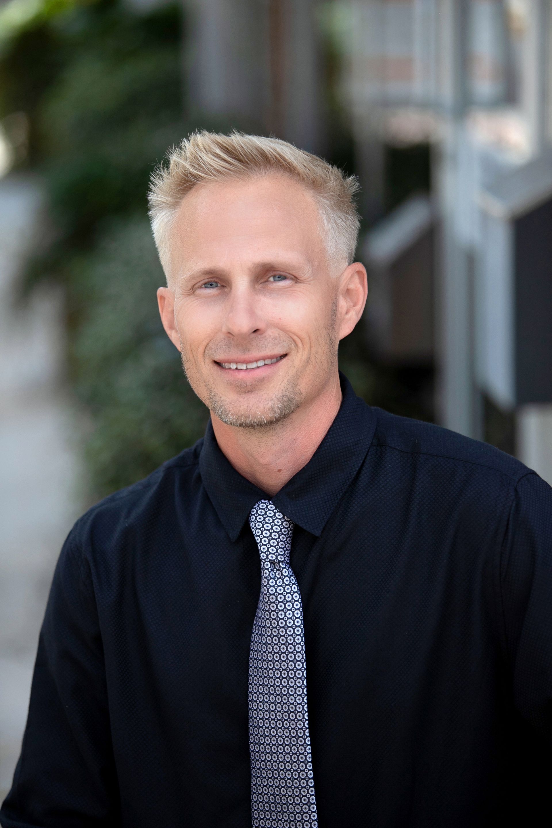 A man in a black shirt and tie is smiling for the camera.