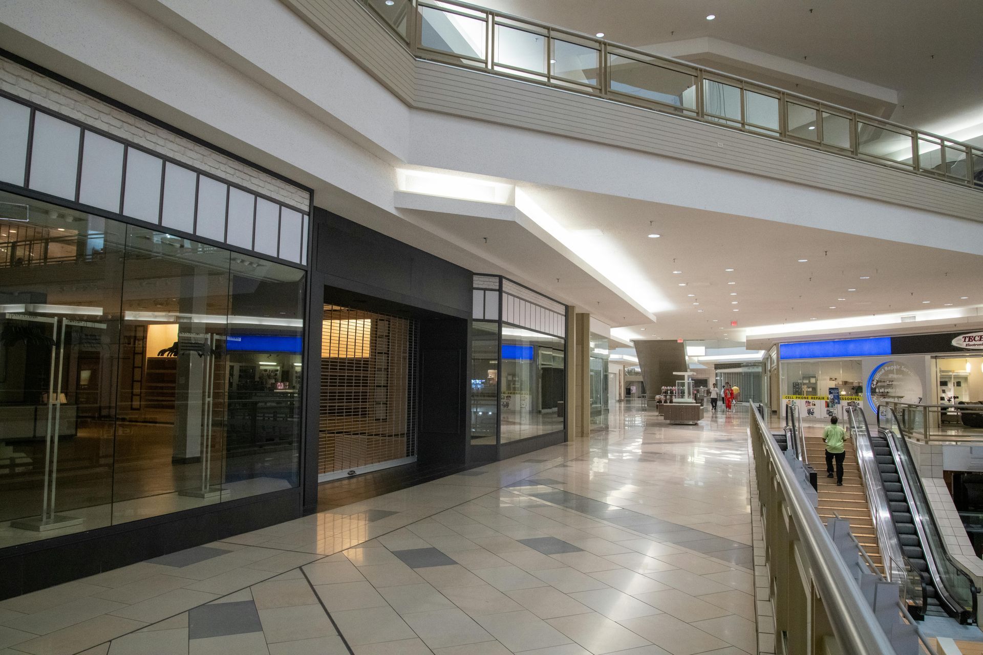 Interior view of a mostly empty mall with closed storefronts, bright lighting, and an escalator.