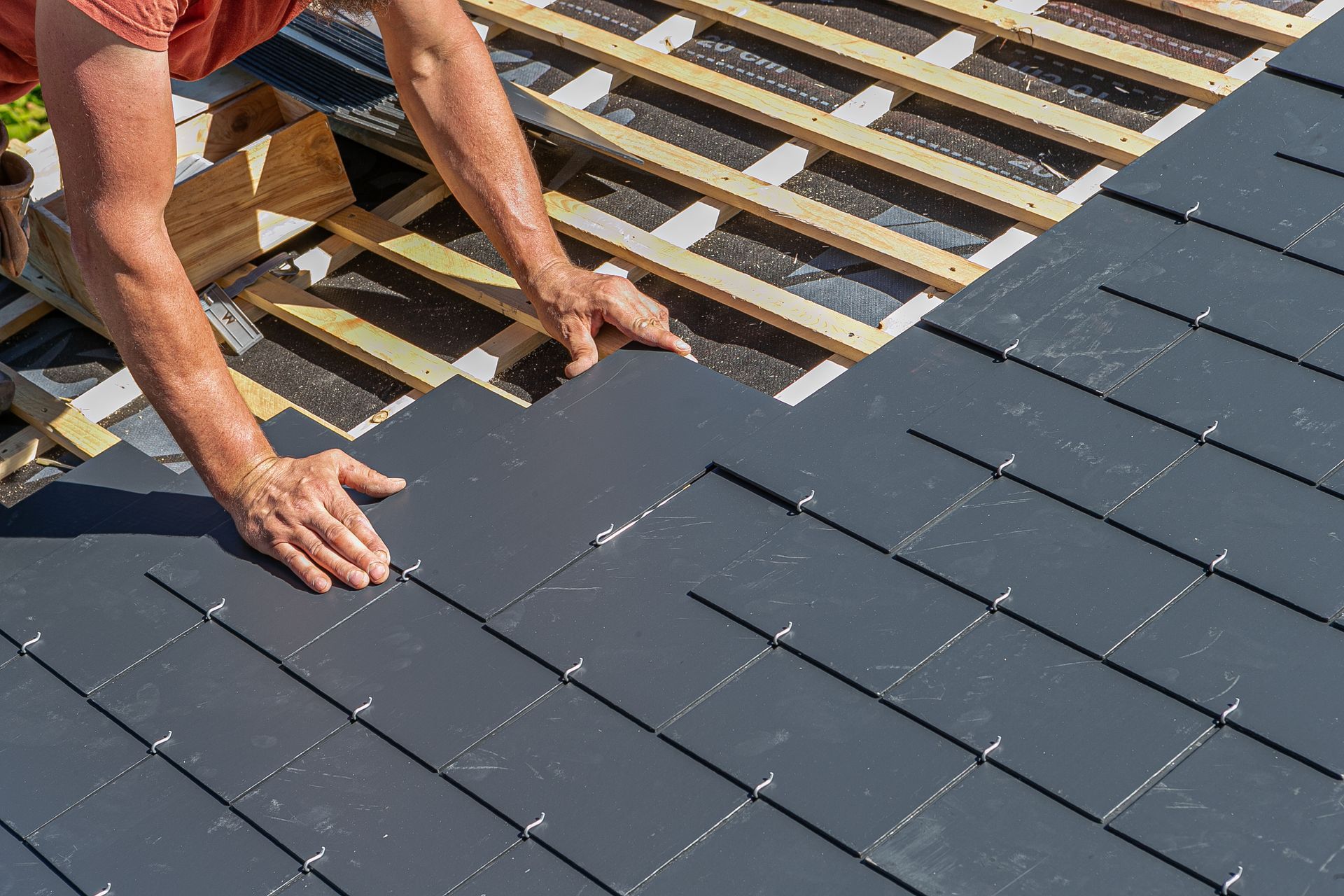 Hands of a roofer installing dark gray roof tiles on a wooden roof.