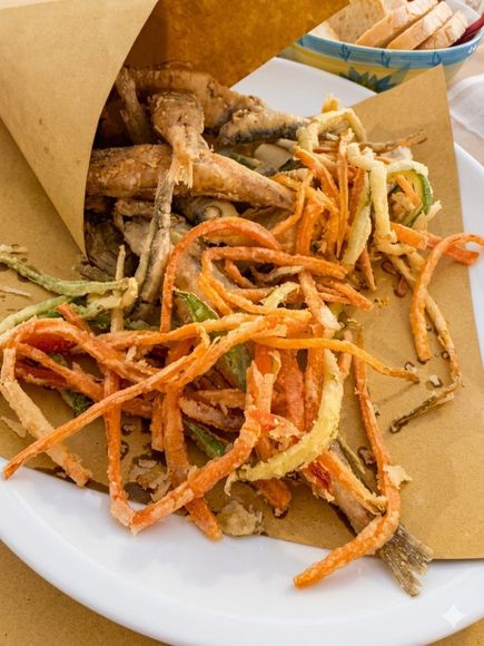 Cono di carta contenente verdure a radice fritte, adagiato su un piatto bianco e guarnito con striscioline croccanti arancioni.