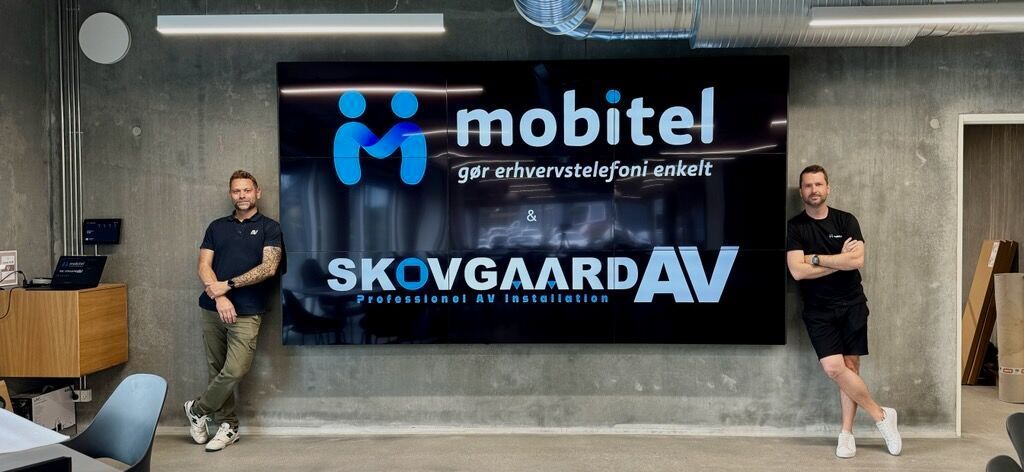 Two men are standing in front of a large videowall that says mobitel skovgaardav.