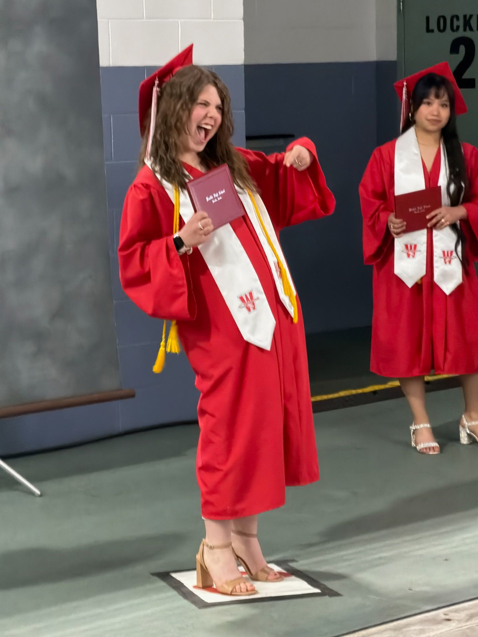 Woman in red graduation gown holding a diploma folder, smiling on a stage beside another graduate.