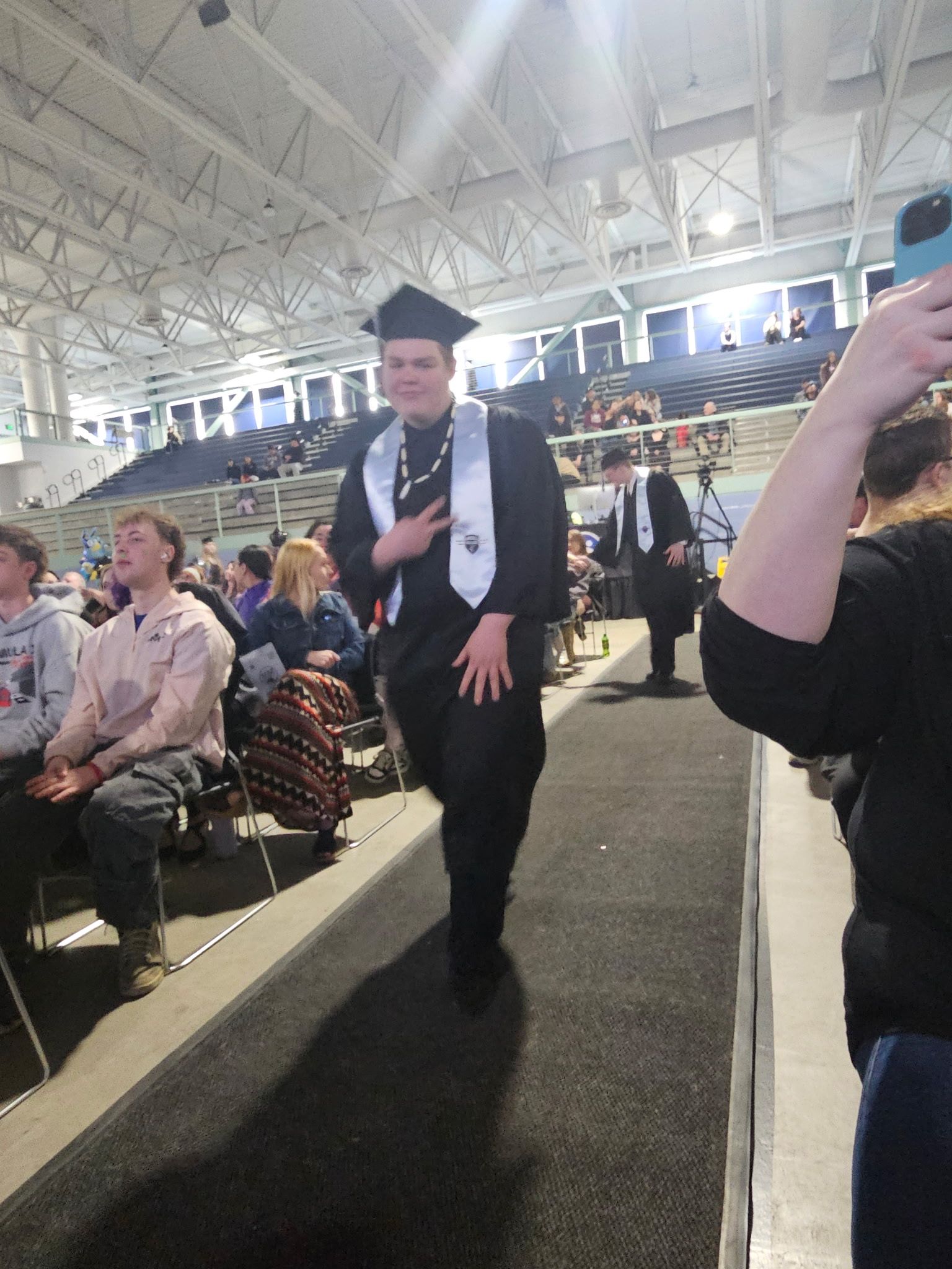Graduate in cap and gown walking down an indoor ceremony aisle, audience seated on both sides.