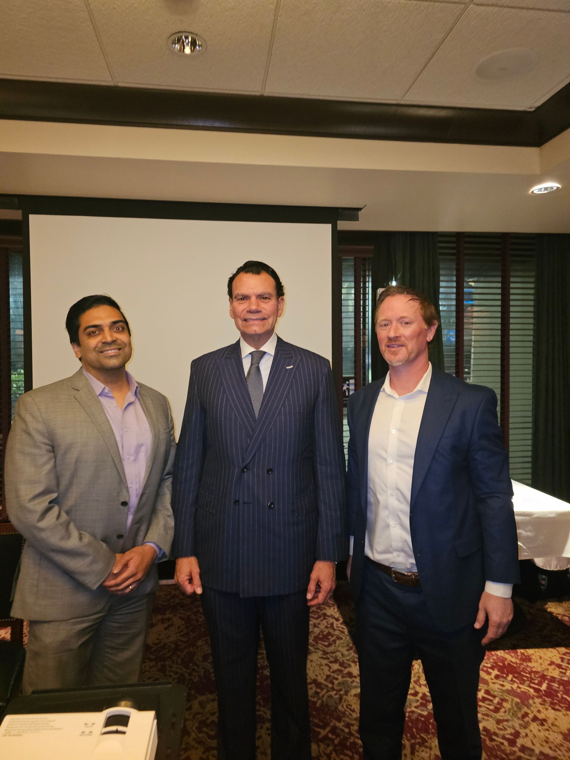 Three men in suits are posing for a picture in front of a projector screen.