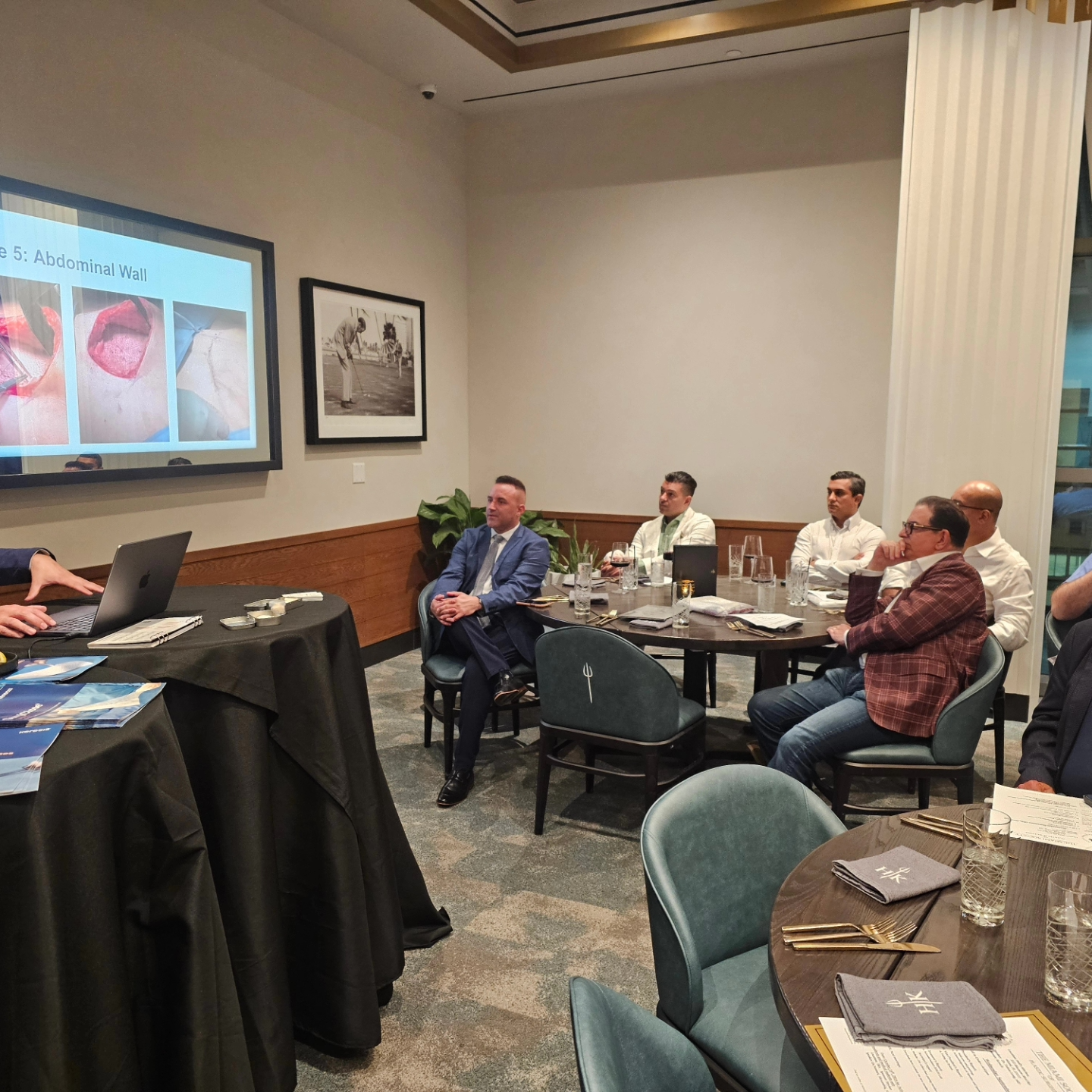 A group of men are sitting at tables in a room with a projector screen.