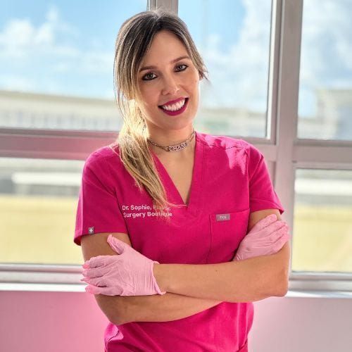 A woman in a pink scrub top and pink gloves is standing in front of a window.