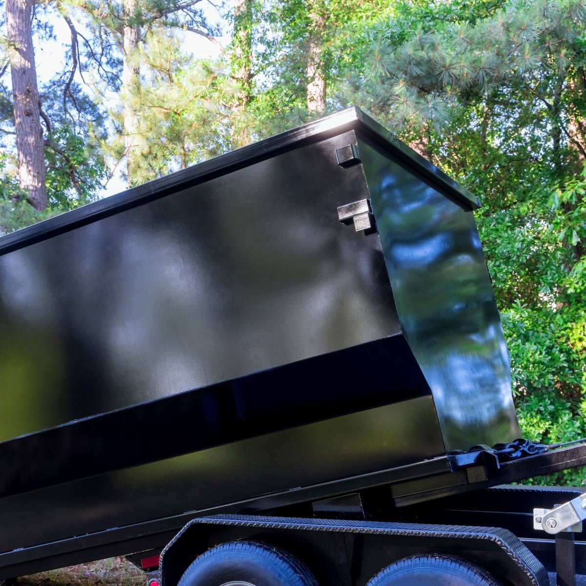 Black dump trailer with raised bed, outdoors with trees in background.