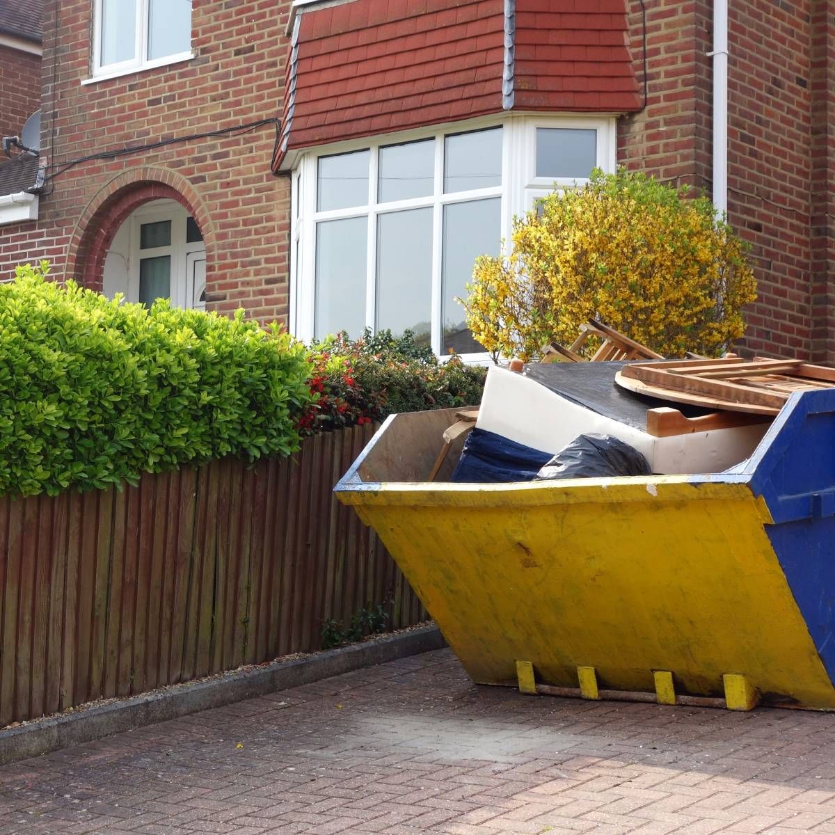 A yellow and blue dumpster overflowing with debris sits on a driveway in front of a brick house.