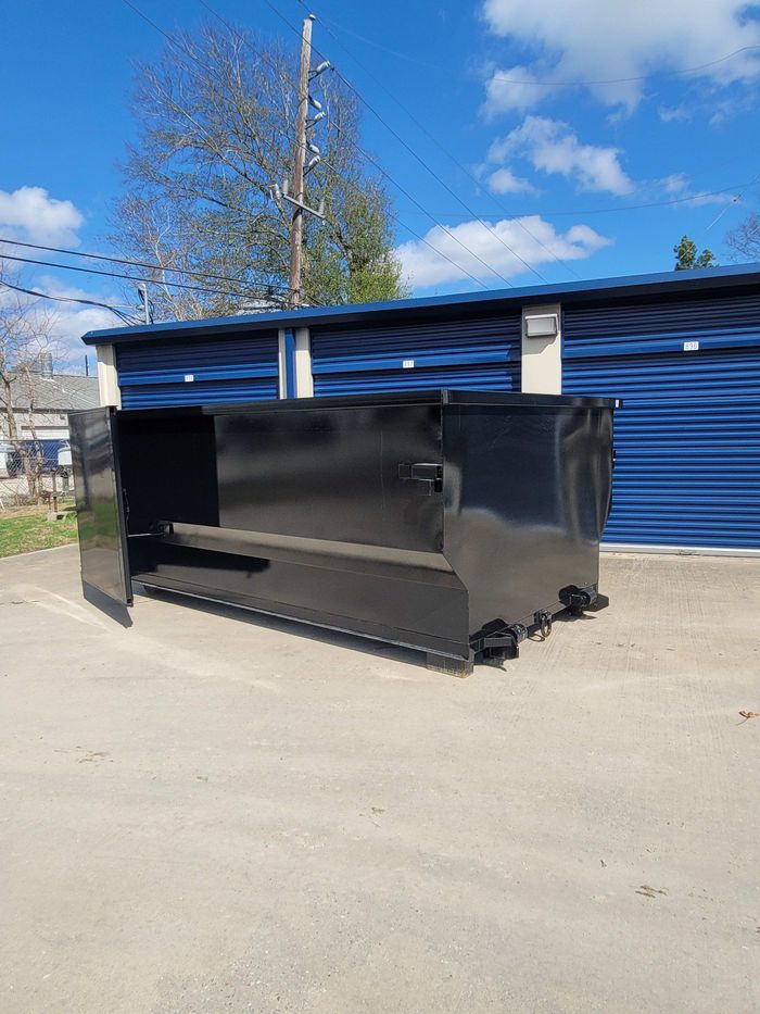 Black dumpster with open door on a paved surface, blue storage units in the background under a partly cloudy sky.