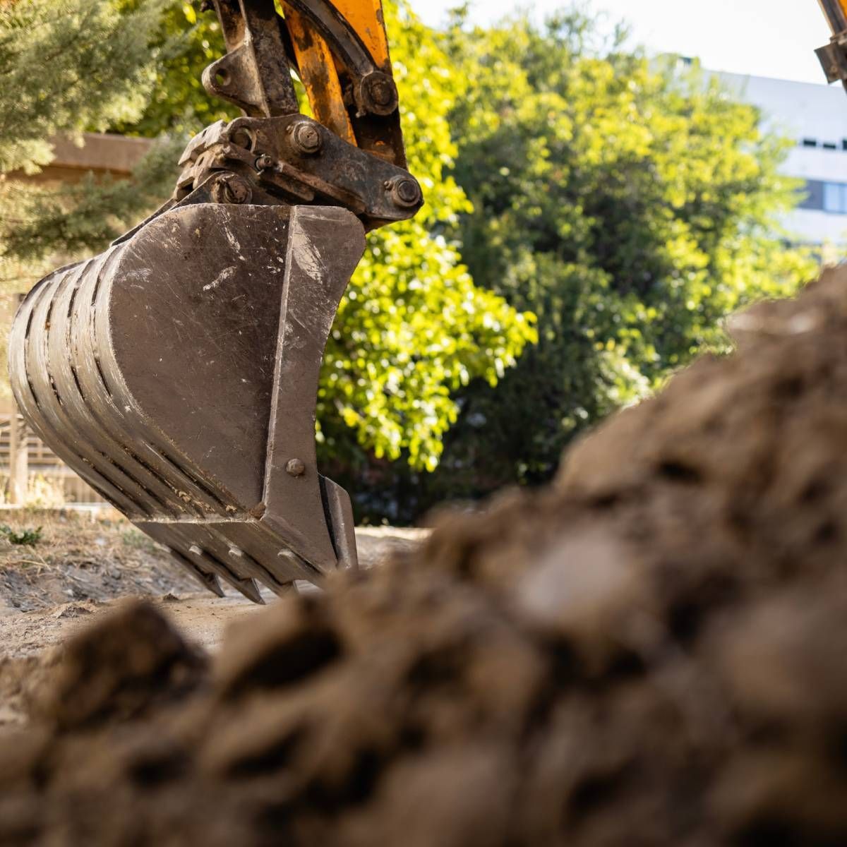 Close-up of an excavator bucket digging into a pile of dirt. Trees and a building are in the blurred background.