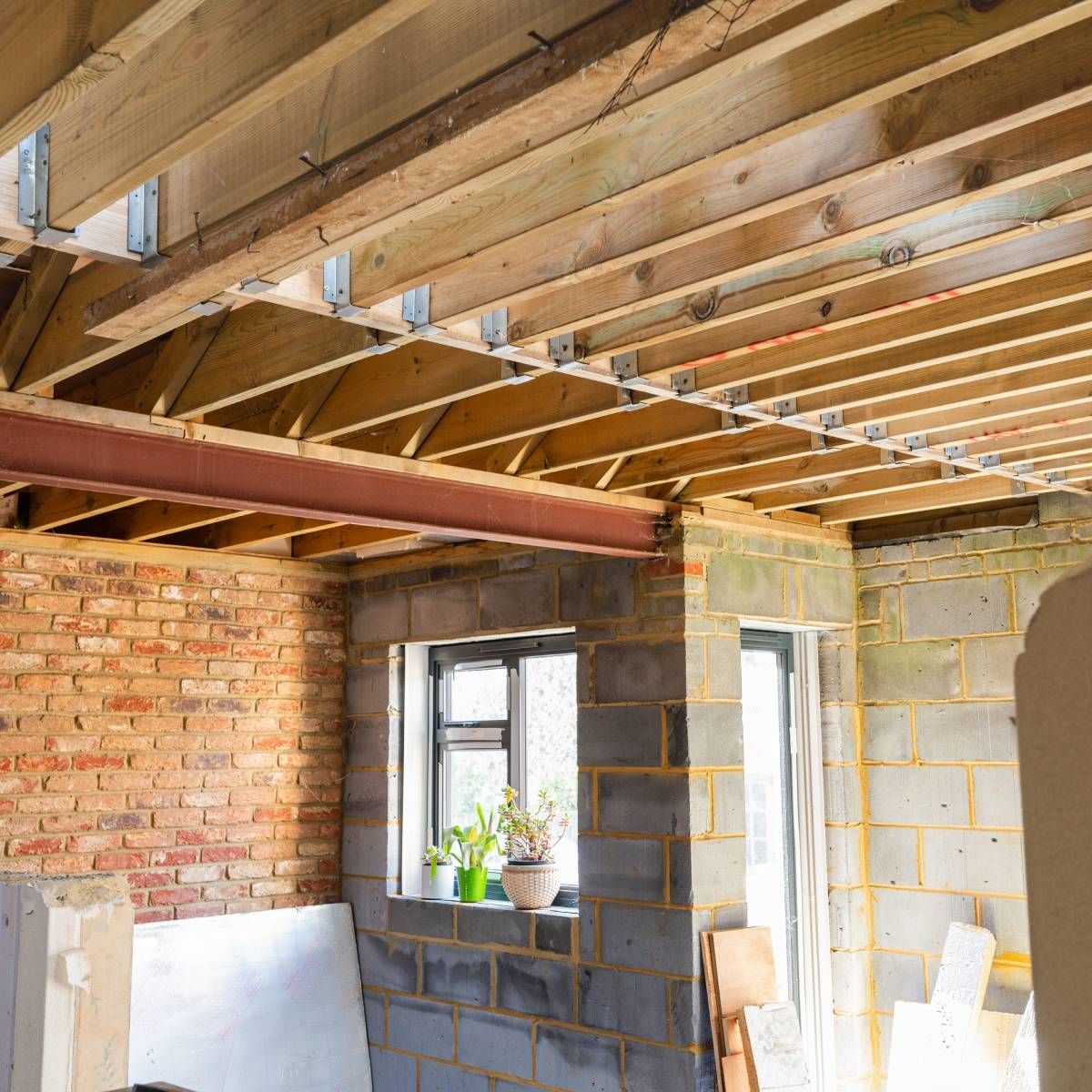 Interior view of a room under construction with exposed wooden joists, steel beam, brick and concrete block walls, and a window.