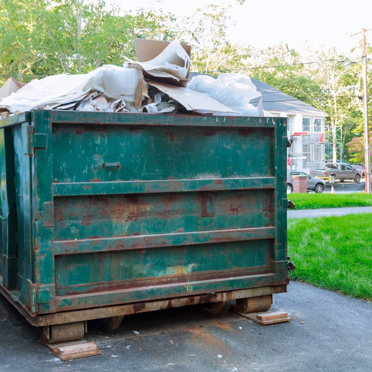 Green dumpster overflowing with construction debris on a driveway in front of a house.