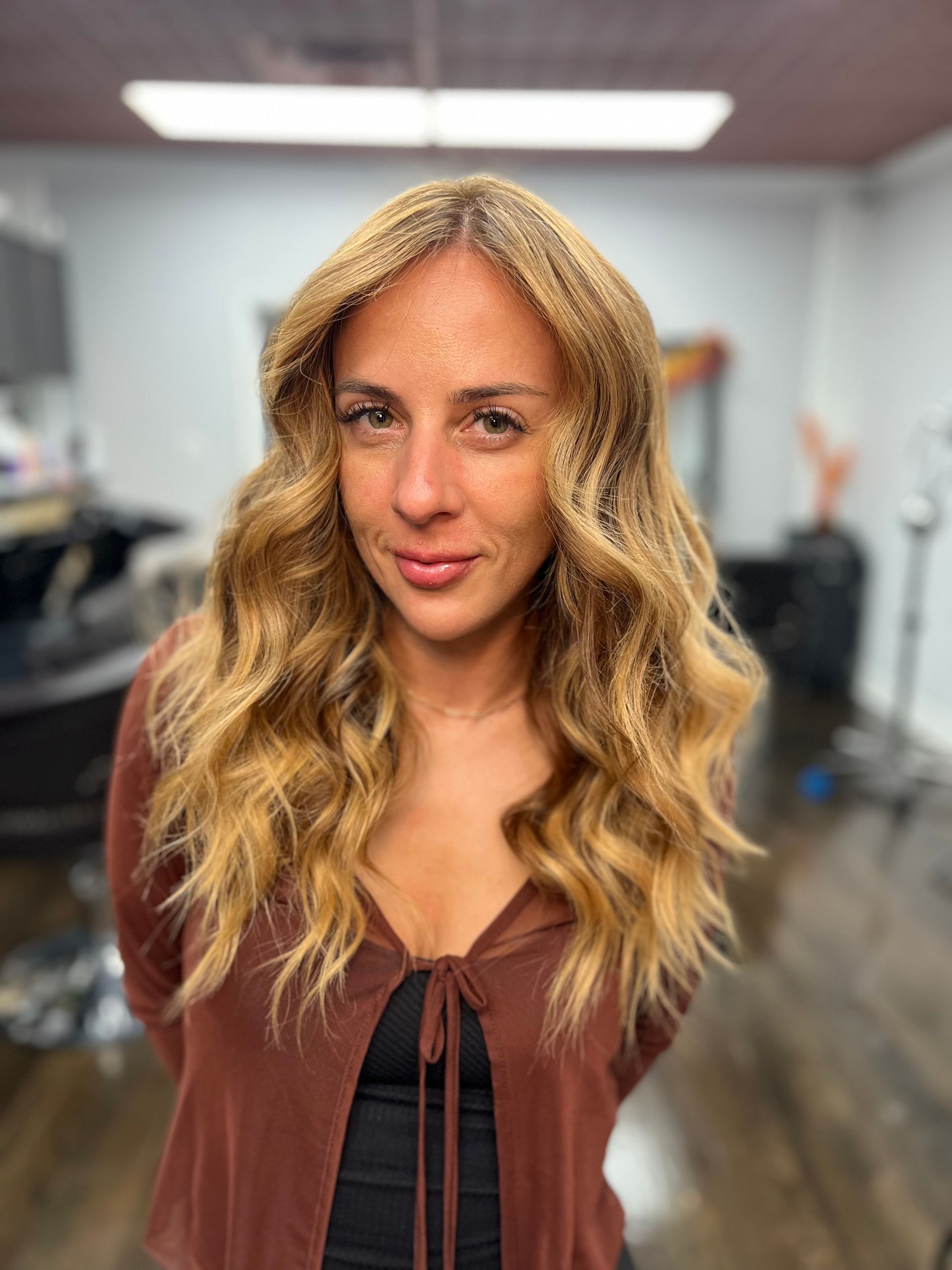 Woman with wavy, blonde hair stands in a salon, wearing a brown cardigan.