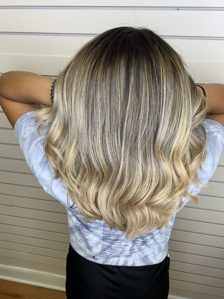 Woman with wavy blonde and brown ombre hair, arms up, against a white wall.