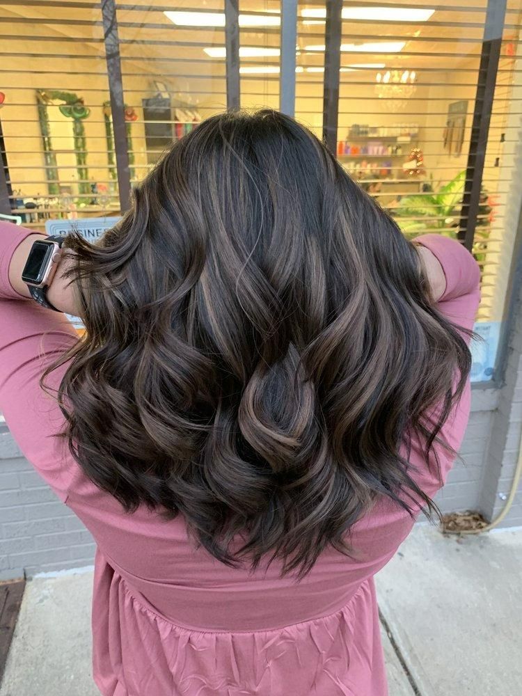 Woman with wavy brown hair and highlights, hands behind head, in front of a shop window.