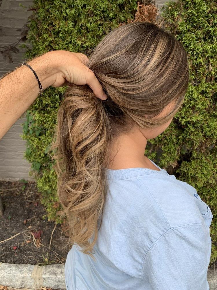 Person with long highlighted hair, pulled into a low ponytail by someone's hand. Green foliage in background.