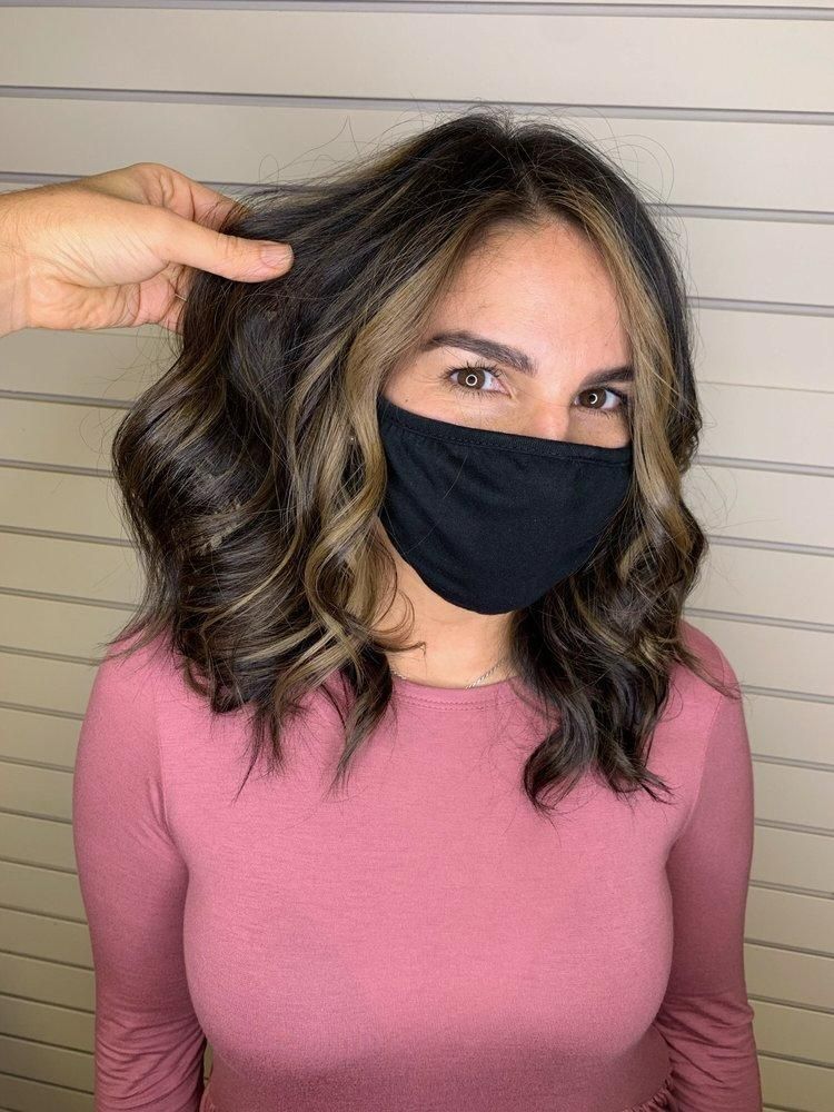 Woman with shoulder-length wavy hair, wearing a black face mask, and pink long-sleeved shirt. Hand touches hair.