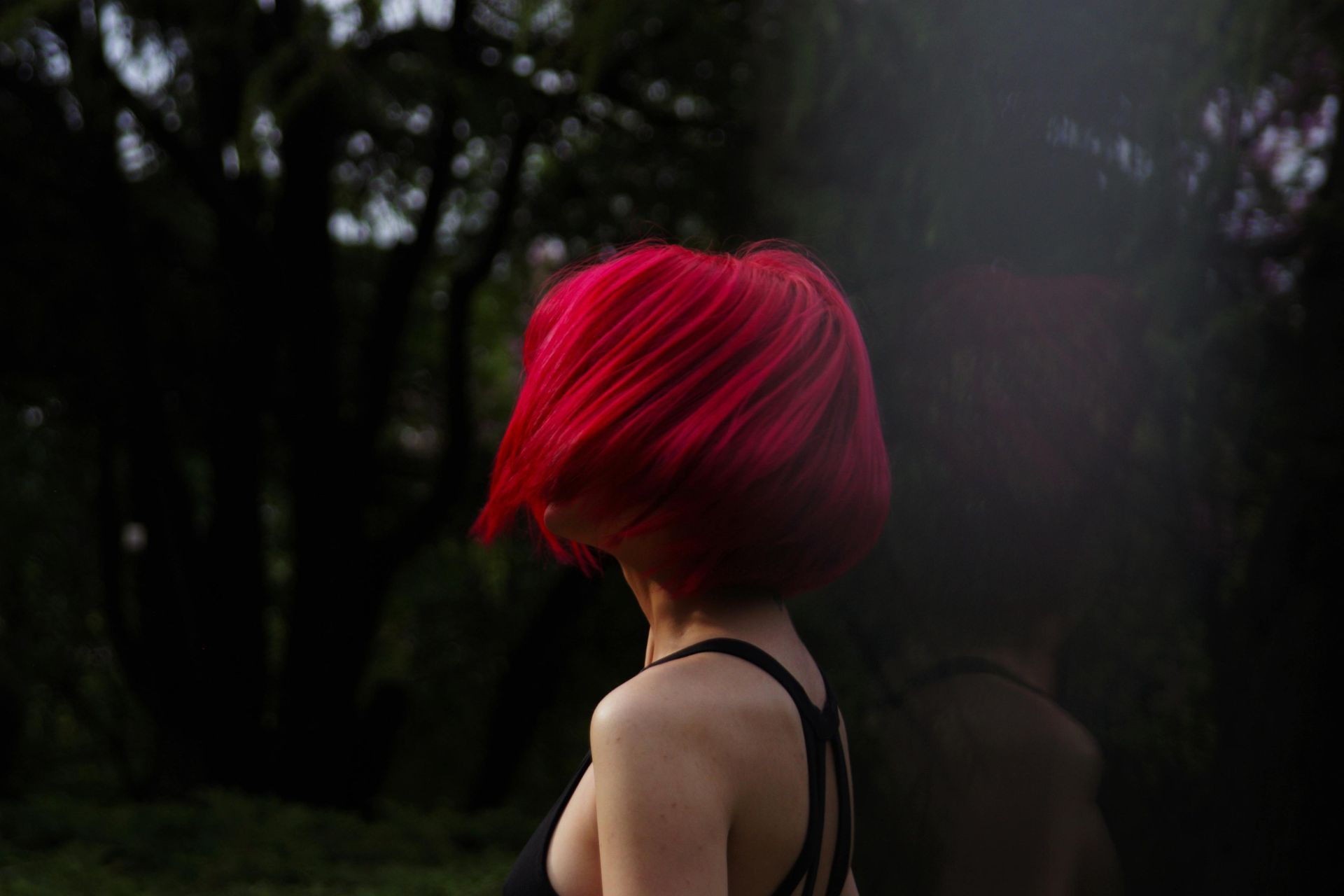 Woman with bright red hair in a dark forest, back to camera, wearing a black top.